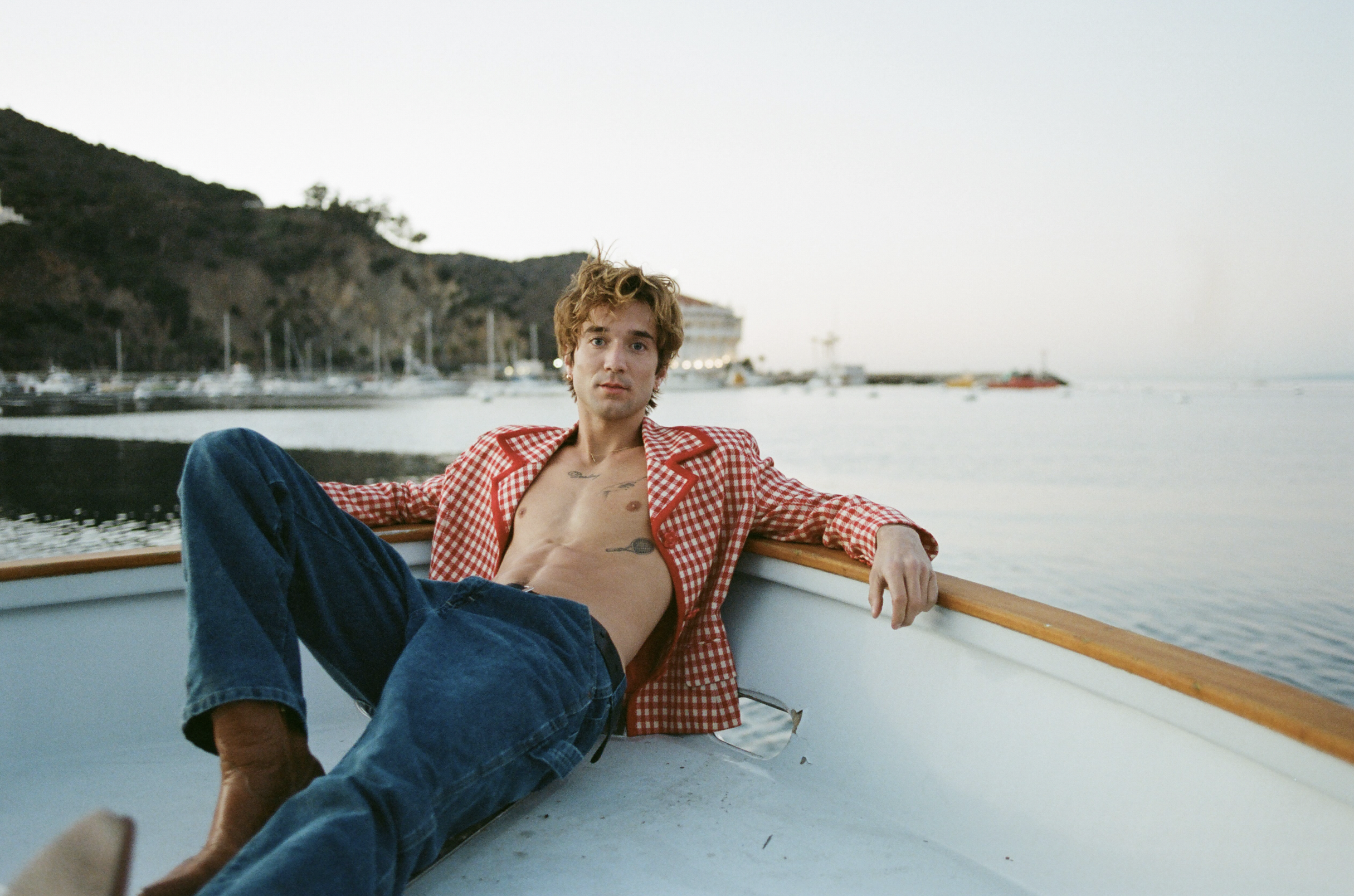 A man with his shirt open laying back on the edge of a boat. He has several tattoos and a serious facial expression.